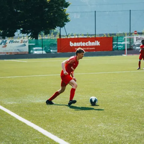 Ein Spieler in roter Trikot führt den Ball auf dem Rasen vor der Werbebande 'bautechnik' beim Spiel Terlan gegen Bruneck.
