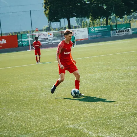 Spieler des AFC Terlan in rotem Trikot führt den Ball auf dem Kunstrasen, im Hintergrund ein zweiter Spieler in Rot und Bandenwerbung.