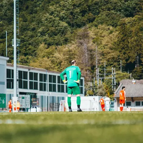Torhüter in grünem Trikot mit Nummer 1 steht von hinten auf dem Rasen und blickt auf das Spielfeld, im Hintergrund Gleisen und Waldhang.
