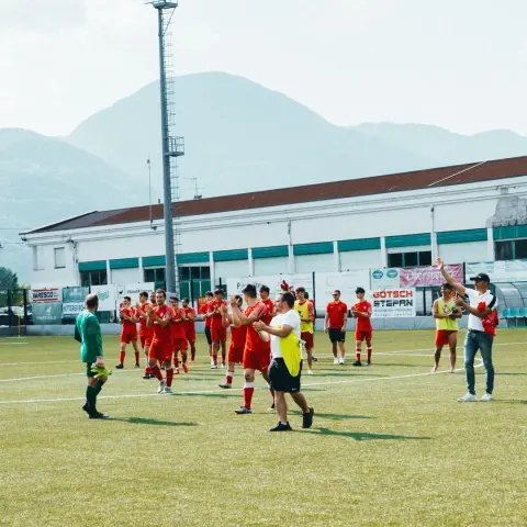 Rot gekleidete Spieler des AFC Terlan klatschen nach dem Spiel gegen Bruneck vor den Fans auf dem Rasenplatz, im Hintergrund Bande und Berge.