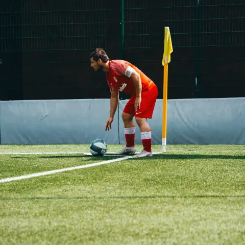 Spieler in roten Trikot mit Kapitänsbinde legt den Ball neben der Eckfahne auf dem Kunstrasen bereit, im Spiel gegen Bruneck.
