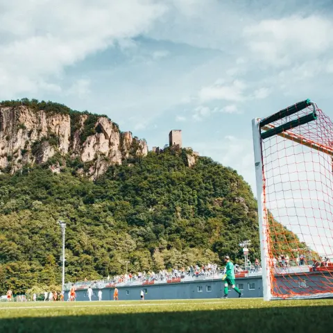 Blick vom Spielfeldrand auf ein Testspiel des AFC Terlan gegen Bruneck, im Vordergrund ein Tor mit orangenem Netz, dahinter Zuschauer und die Burgruine auf dem Felsen.