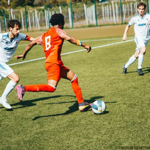 Spielszene: Spieler in Orange mit Nummer 8 zieht den Ball am Gegner in Weiß vorbei, ein weiterer Weißer läuft im Hintergrund mit.
