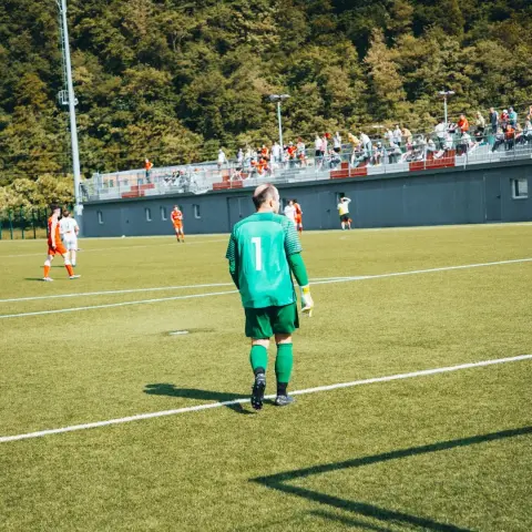 Torhüter in grünem Trikot mit der Nummer 1 läuft über den Rasenplatz, im Hintergrund Spieler in Orange und Weiß und Zuschauer auf der Tribüne.