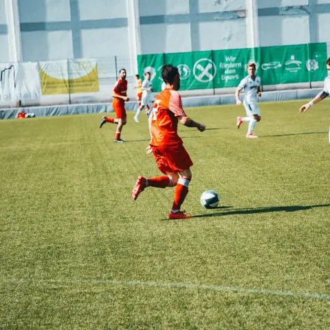 Spielszene: Ein Spieler in roter Trikot führt den Ball am Rand eines Kunstrasenplatzes, im Hintergrund weiße Gegner und grüne Bandenwerbung.