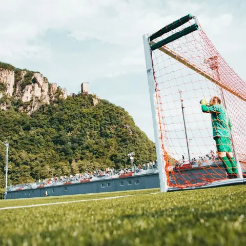 Torwart in grünem Trikot steht im orangen Netz des Tors auf dem Kunstrasen, im Hintergrund Zuschauer auf der Tribüne und der Felsen mit Burgruine.
