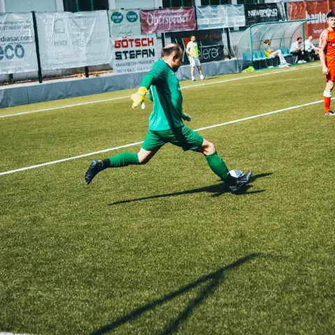 Torhüter in grünem Trikot schlägt den Ball mit dem Fuß auf dem Kunstrasen, im Hintergrund Bandenwerbung und Spieler in Orange.