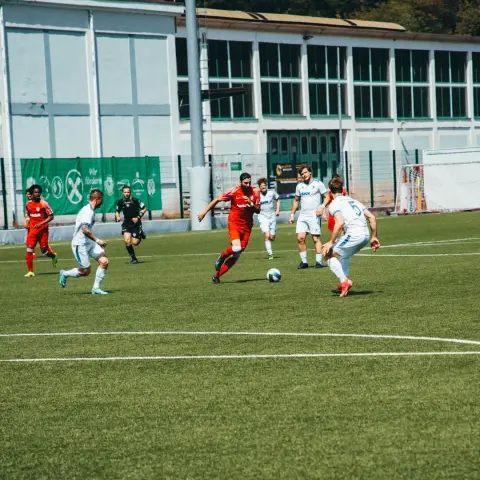 Spielszene: Ein Spieler in Rot dribbelt den Ball auf dem Rasen, umringt von Gegenspielern in Weiß beim Spiel AFC Terlan gegen Bruneck.