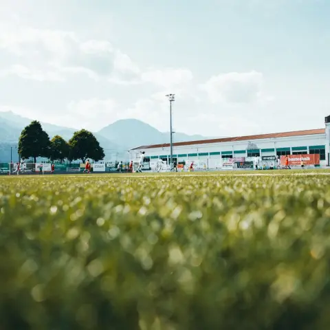 Spielszene auf dem Rasen des Sportplatzes Terlan mit Spielern in Rot und Weiß, im Hintergrund Tribüne mit Bandenwerbung und Bergkulisse.