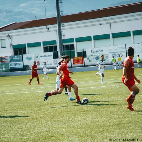 Rot gekleideter Spieler des AFC Terlan führt den Ball im Spiel gegen Bruneck, Gegner in Weiß verfolgt ihn auf dem Kunstrasen.