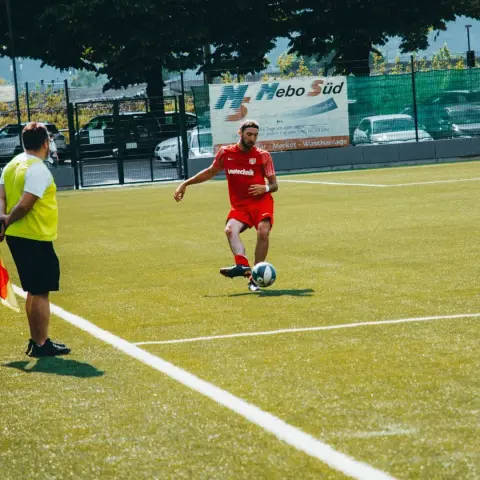 Spieler in roter Bautechnik-Trikot führt bei einem Spiel gegen Bruneck den Ball auf dem Kunstrasen, im Hintergrund Bande mit Mebo Süd-Werbung.