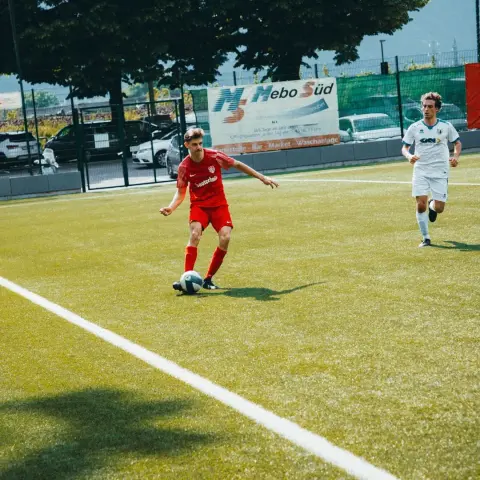 Spielszene: Ein Spieler in roter Trikot führt den Ball am Rand des Kunstrasenplatzes, ein Gegner in Weiß läuft hinterher, im Hintergrund Bande und Bäume.