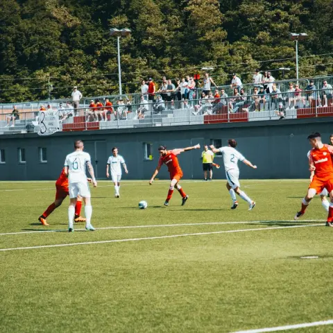 Spielszene: Ein Spieler in Rot führt den Ball auf dem Rasen, während Spieler in Weiß und Rot um ihn kämpfen, im Hintergrund Zuschauer auf der Tribüne.