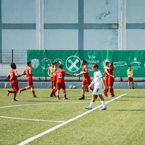 Spieler des AFC Terlan in Rot feiern vor einer grünen Werbebande mit Sponsorenlogos, ein Gegenspieler in Weiß geht vorbei - Spiel gegen Bruneck.