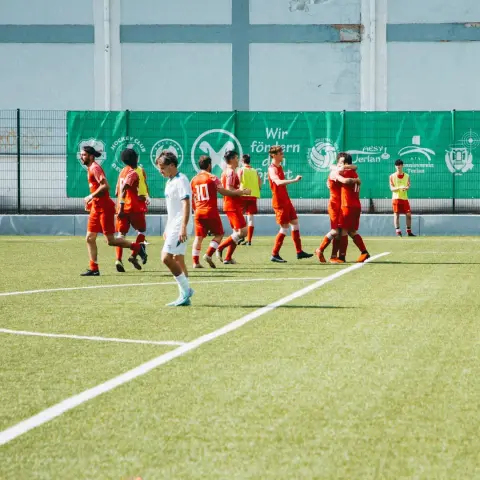 Spieler des AFC Terlan in Rot jubeln auf dem Kunstrasen vor einer grünen Bandenwerbung, während ein Gegenspieler in Weiß enttäuscht vorbeigeht.