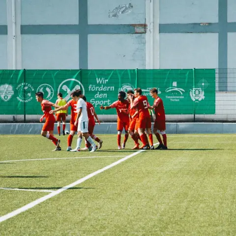 Spieler des AFC Terlan in roten Trikots jubeln auf dem Platz nach einem Tor, ein Gegner in Weiß steht daneben, im Hintergrund Werbebande mit Sponsoren.