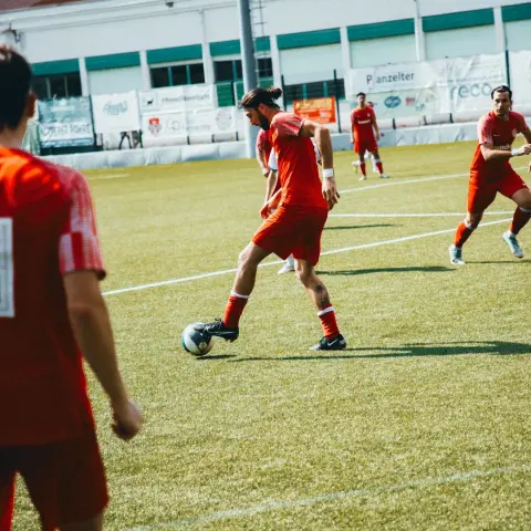 Spieler des AFC Terlan in roter Trikot führt den Ball auf dem Kunstrasen während des Spiels gegen Bruneck, im Hintergrund Bandenwerbung.