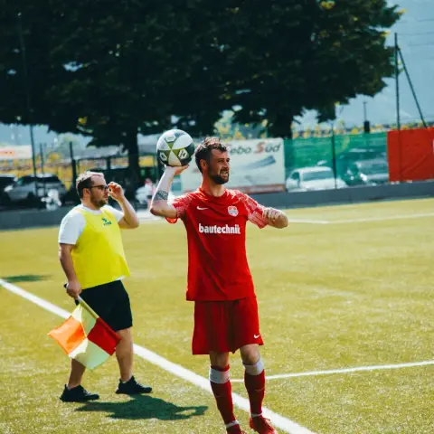 Spieler in roter bautechnik-Trikot wirft den Ball am Spielfeldrand ein, daneben steht ein Linienrichter mit gelber Weste und Fahne.