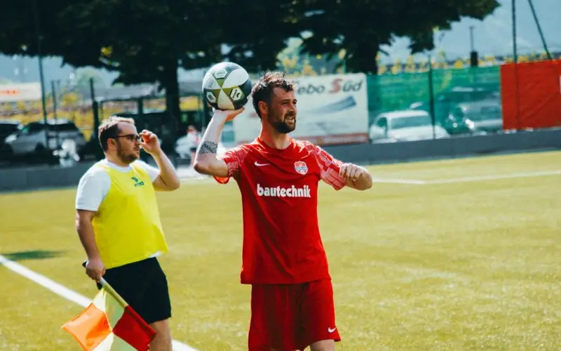 Spieler in roter bautechnik-Trikot wirft den Ball am Spielfeldrand ein, daneben steht ein Linienrichter mit gelber Weste und Fahne.
