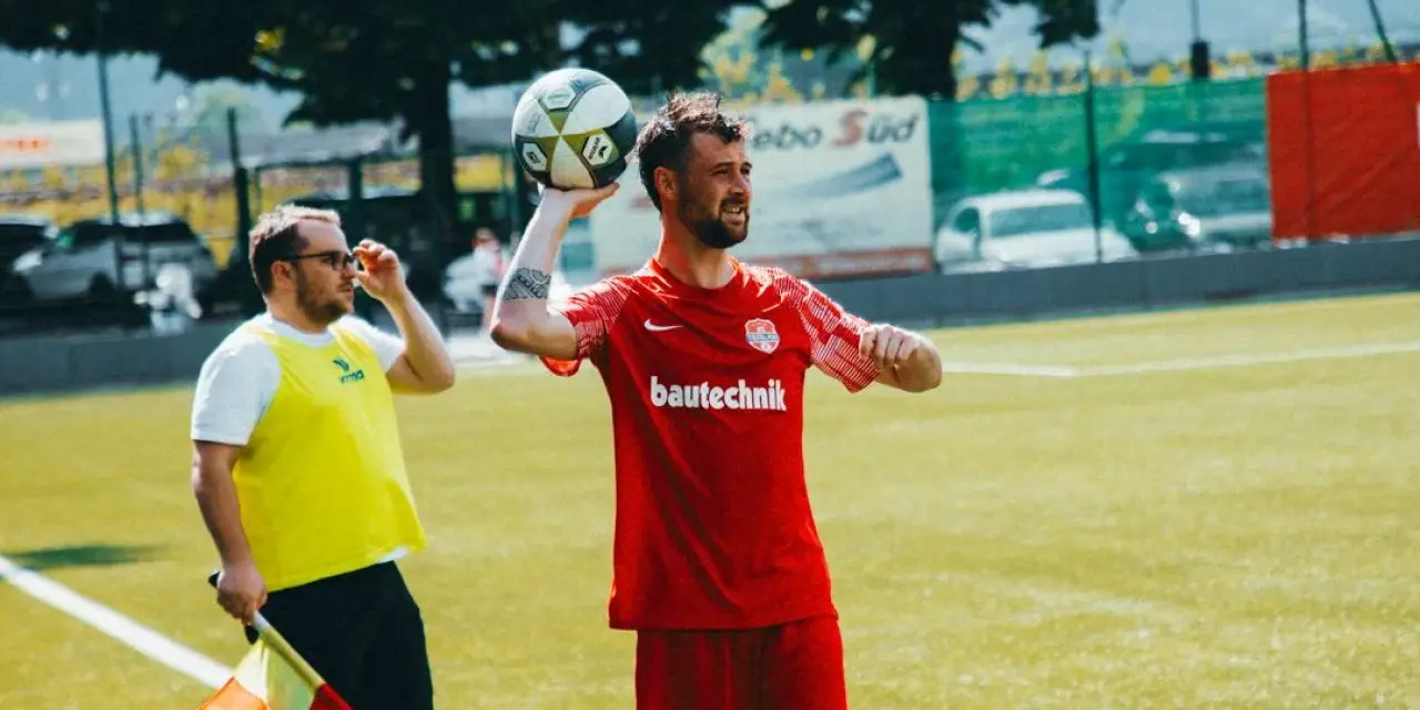 Spieler in roter bautechnik-Trikot wirft den Ball am Spielfeldrand ein, daneben steht ein Linienrichter mit gelber Weste und Fahne.