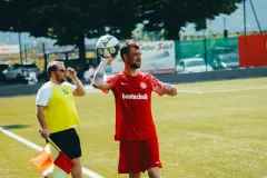 Spieler in roter bautechnik-Trikot wirft den Ball am Spielfeldrand ein, daneben steht ein Linienrichter mit gelber Weste und Fahne.