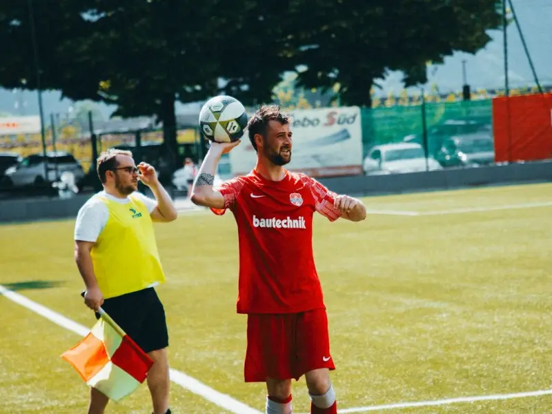 Spieler in roter bautechnik-Trikot wirft den Ball am Spielfeldrand ein, daneben steht ein Linienrichter mit gelber Weste und Fahne.