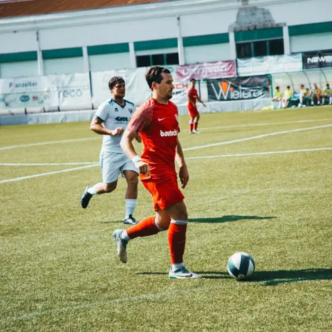 Spieler des AFC Terlan in orangem Trikot führt den Ball auf dem Rasen, verfolgt von einem Gegenspieler in Weiß beim Spiel gegen Bruneck.
