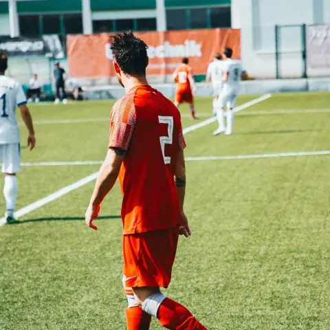 Spieler des AFC Terlan in orangerotem Trikot mit Nummer 2 geht auf dem Rasen, im Hintergrund Spieler in Weiß und Werbebande Bautechnik.