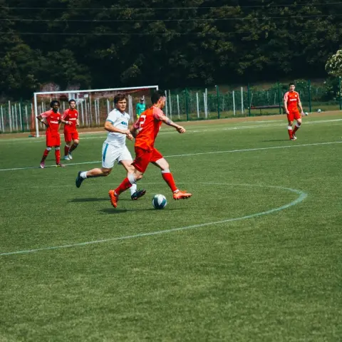 Spielszene: Ein Spieler in Rot führt den Ball am Mittelkreis, während ein Gegner in Weiß von hinten nachsetzt, im Hintergrund weitere Spieler in Rot.