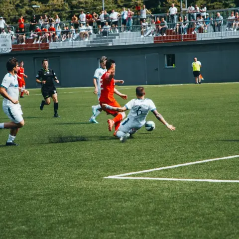 Spielszene: Ein Spieler in Weiß grätscht im Zweikampf gegen einen Gegner in Orange, der Ball rollt auf dem Rasen des Stadions.