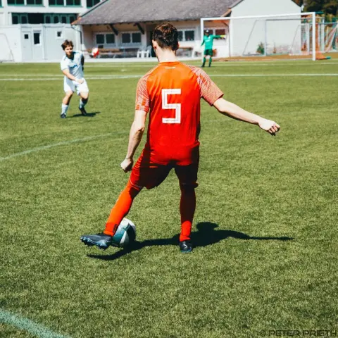 Spieler in orangem Trikot mit Nummer 5 führt den Ball am Fuß über den Rasen, im Hintergrund ein Gegenspieler in Weiß und der Torhüter in Grün.