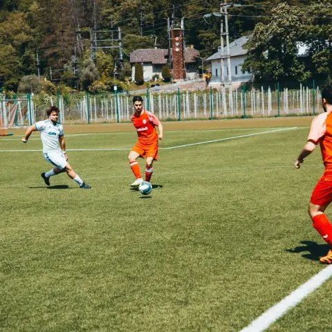 Spielszene auf einem Kunstrasenplatz: Ein Spieler in Orange am Ball wird von einem Gegner in Weiß verfolgt, im Hintergrund Bäume und Gebäude.