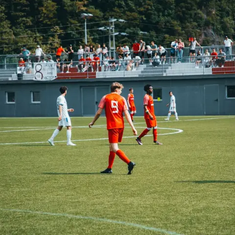 Spielszene: Spieler in Orange mit Trikotnummer 9 geht über den Rascher, im Hintergrund Zuschauer auf der Tribüne und ein Banner mit Sponsorenschrift.