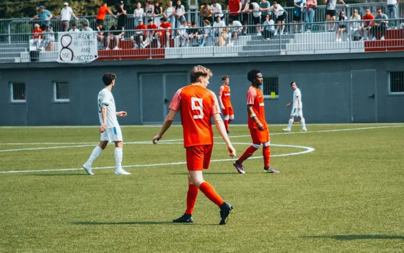 Spielszene: Spieler in Orange mit Trikotnummer 9 geht über den Rascher, im Hintergrund Zuschauer auf der Tribüne und ein Banner mit Sponsorenschrift.