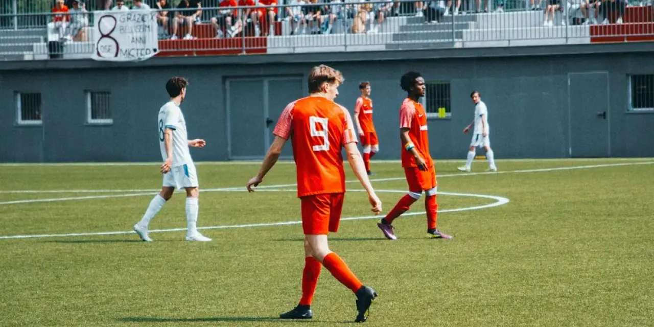 Spielszene: Spieler in Orange mit Trikotnummer 9 geht über den Rascher, im Hintergrund Zuschauer auf der Tribüne und ein Banner mit Sponsorenschrift.