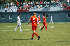 Spielszene: Spieler in Orange mit Trikotnummer 9 geht über den Rascher, im Hintergrund Zuschauer auf der Tribüne und ein Banner mit Sponsorenschrift.
