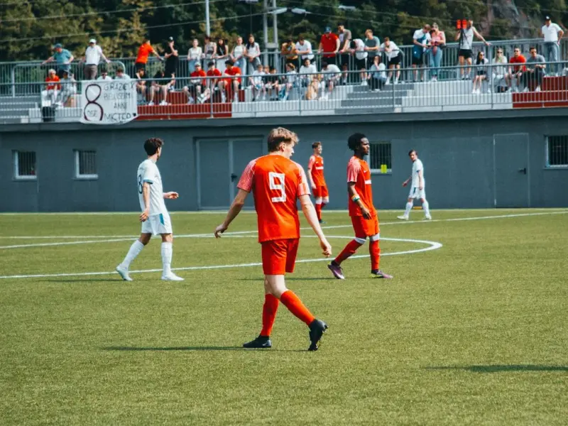 Spielszene: Spieler in Orange mit Trikotnummer 9 geht über den Rascher, im Hintergrund Zuschauer auf der Tribüne und ein Banner mit Sponsorenschrift.