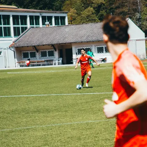 Spieler des AFC Terlan in roter Trikot führt den Ball auf dem Rasen, im Vordergrund ein Mitspieler von hinten, im Hintergrund das Vereinsgelände.