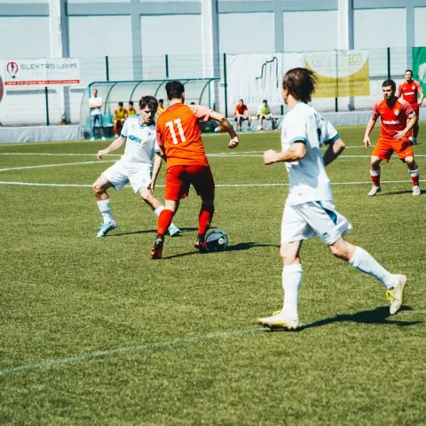 Spielszene: Ein Spieler in Orange mit Nummer 11 führt den Ball gegen einen Gegner in Weiß, im Hintergrund stehen weitere Spieler.