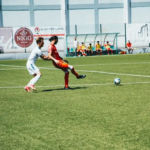 Spielszene: Ein Spieler in Rot wird auf dem Kunstrasen von einem Gegner in Weiß bedrängt, während der Ball im Vordergrund rollt.