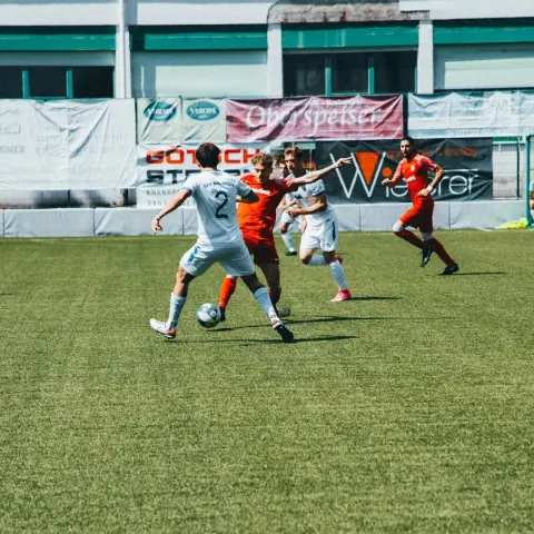 Spielszene: Ein Spieler in Weiß mit Trikotnummer 2 führt den Ball gegen einen Gegner in Rot, im Hintergrund Banden und Mitspieler.