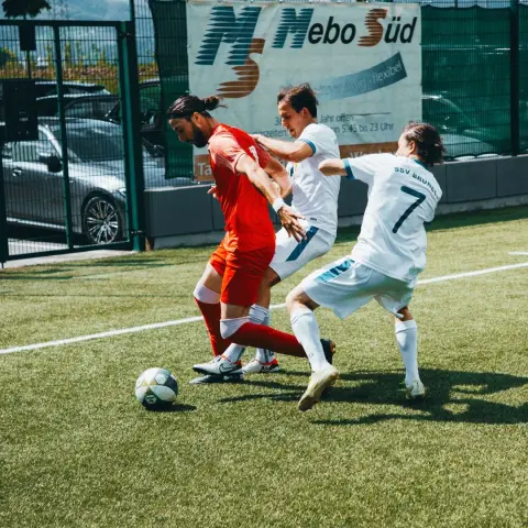 Spielszene: Ein Spieler in Rot wird im Zweikampf von zwei Spielern des SSV Bruneck in Weiß um den Ball bedrängt, im Hintergrund eine Bande mit Sponsor Mebo Süd.