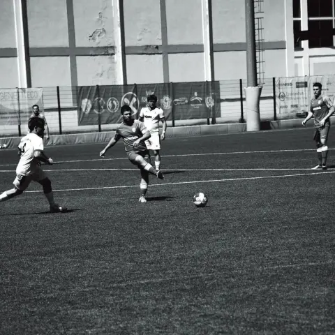 Schwarz-weiße Spielszene: Ein Spieler in grauem Trikot zieht den Ball am Boden, daneben ein Gegenspieler in Weiß bei einem Spiel gegen Bruneck.