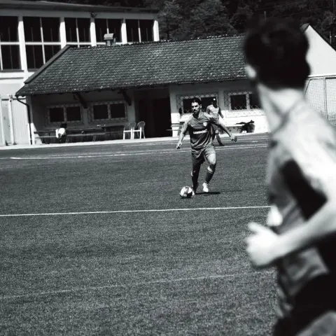 Spieler im Trikot mit Aufschrift führt in Schwarzweiß den Ball über einen Rasenplatz, im Vordergrund unscharf ein weiterer Spieler, im Hintergrund das Vereinsgebäude.