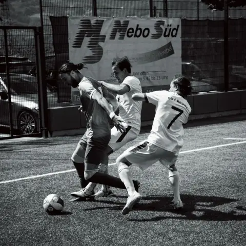 Schwarzweißfoto einer Spielszene: Drei Spieler kämpfen um den Ball, rechts ein Spieler des SSV Bruneck mit Trikot Nr. 7, im Hintergrund eine Werbebande von Nebo Süd.