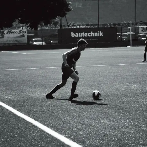 Schwarz-weißes Foto: Ein Spieler des AFC Terlan führt den Ball über den Kunstrasen, im Hintergrund die Bande mit dem Schriftzug bautechnik.