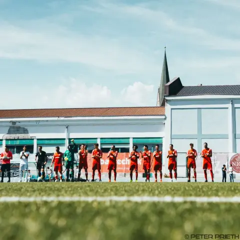 Spieler in roten Trikots stehen am Spielfeldrand Spalier und klatschen, daneben Schiedsrichter und Gegner in Weiß; im Hintergrund Bandenwerbung und Kirchturm.