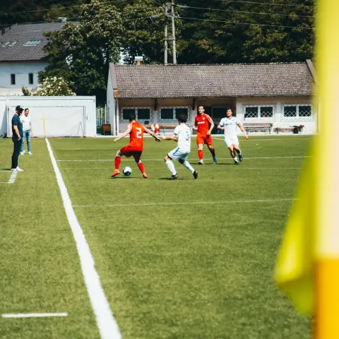 Spielszene: Ein Spieler in Orange mit der Nummer 2 führt den Ball gegen zwei Gegner in Weiß auf dem Rasenplatz, im Hintergrund Tribüne und Bäume.