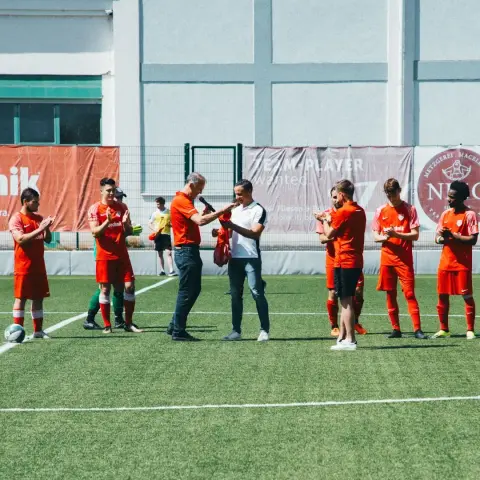 Spieler des AFC Terlan in Orange klatschen auf dem Rasenplatz, während zwei Männer in der Mitte ein Trikot übergeben und dabei ins Mikrofon sprechen.