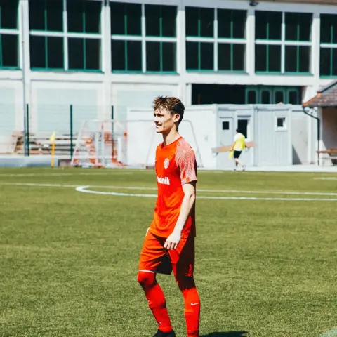 Ein Fußballspieler in oranger Trikotkombination mit Sponsorenaufdruck steht lächelnd auf dem Kunstrasen, im Hintergrund ein Schulgebäude.
